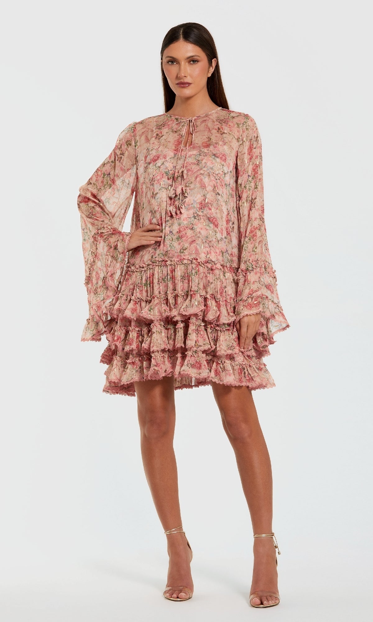 A woman poses in the Mac Duggal 8336 Print Short Wedding Guest Dress with long sleeves, pink floral pattern, ruffled layers, and beige high-heeled sandals against a plain light background.